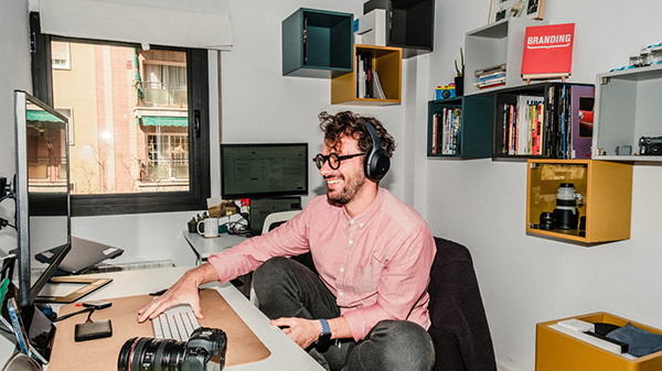 Man working from home at desk with headphones on, smiling while using a computer in a modern home office.